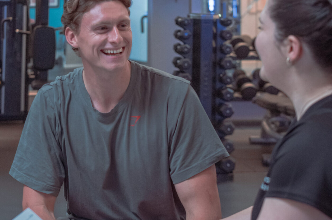 A man sits at a table on the gym floor, smiling as he speaks with a fitness advisor during a consultation. The advisor holds a clipboard and pen.